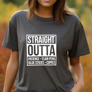 May include: Dark gray t-shirt with the words "STRAIGHT OUTTA" in a white box, followed by "PATIENCE &bull; FLAIR PENS &bull; GLUE STICKS &bull; COPIES" in a white font. The shirt is short-sleeved.