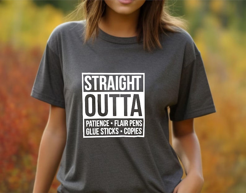 May include: Dark gray t-shirt with the words "STRAIGHT OUTTA" in a white box, followed by "PATIENCE &bull; FLAIR PENS &bull; GLUE STICKS &bull; COPIES" in a white font. The shirt is short-sleeved.
