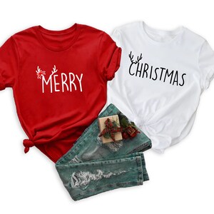 May include: Two short-sleeved t-shirts, one red and one white, with Christmas-themed text. The red shirt says "MERRY" with antler and bow accents. The white shirt says "CHRISTMAS" with antler accents. Blue jeans and a small gift box are also in the image.