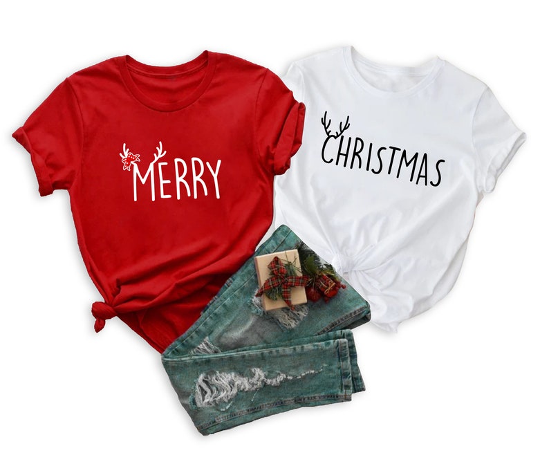 May include: Two short-sleeved t-shirts, one red and one white, with Christmas-themed text. The red shirt says "MERRY" with antler and bow accents. The white shirt says "CHRISTMAS" with antler accents. Blue jeans and a small gift box are also in the image.