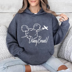 Puede incluir: Una sudadera con capucha azul oscuro con un gr&aacute;fico blanco de la cabeza de Mickey Mouse en nubes con un avi&oacute;n volando sobre ella. El texto "Disney Bound!" est&aacute; debajo del gr&aacute;fico.