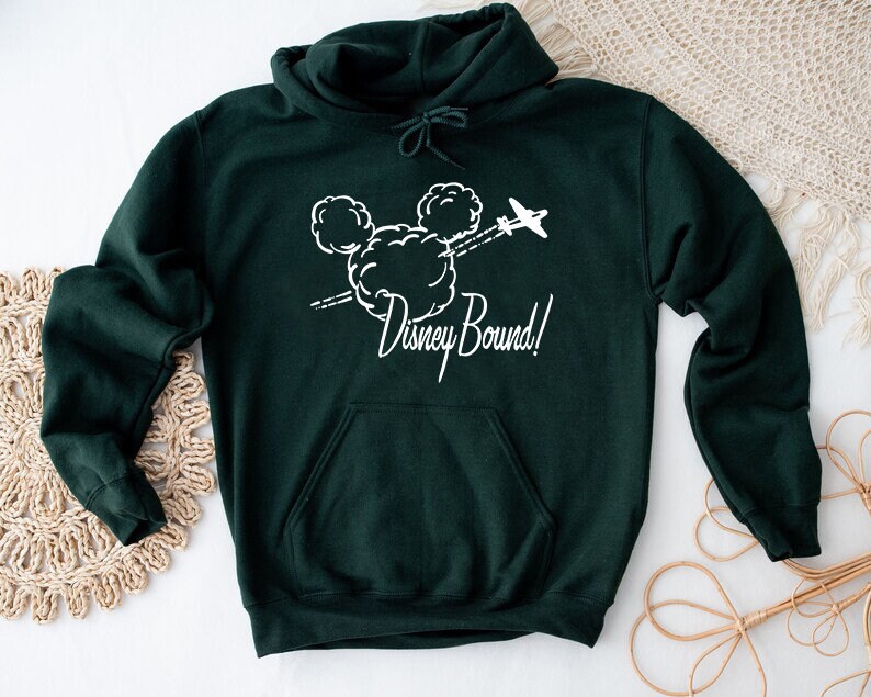 Puede incluir: Una sudadera con capucha verde oscuro con un gr&aacute;fico blanco de la cabeza de Mickey Mouse en nubes con un avi&oacute;n volando por encima. El texto "Disney Bound!" est&aacute; debajo del gr&aacute;fico.