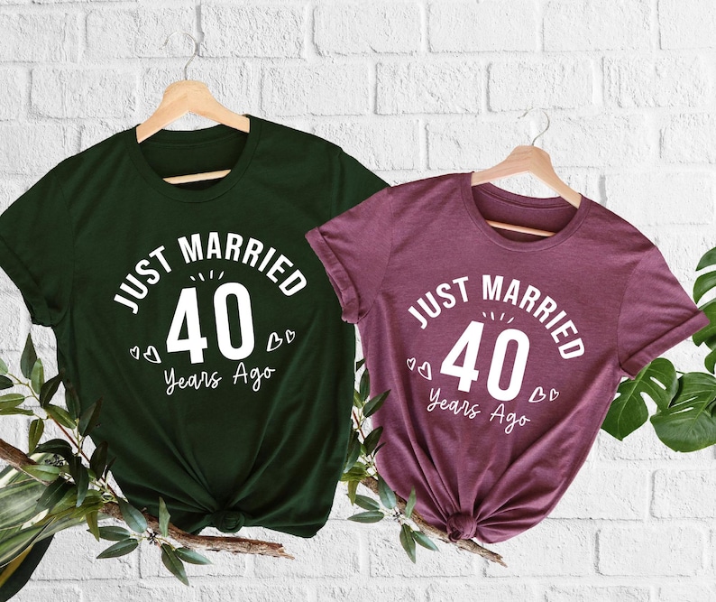 May include: Two short-sleeved t-shirts, one dark green and one burgundy, hang on wooden hangers. Each shirt features the white text "Just Married 40 Years Ago" with a stylized "40" in the center. The shirts are displayed against a white brick wall.