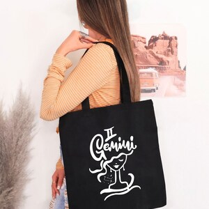 May include: A black tote bag with a white graphic of a woman's face and the text "Gemini" above it. The woman's face is stylized and has a constellation of stars around her head.