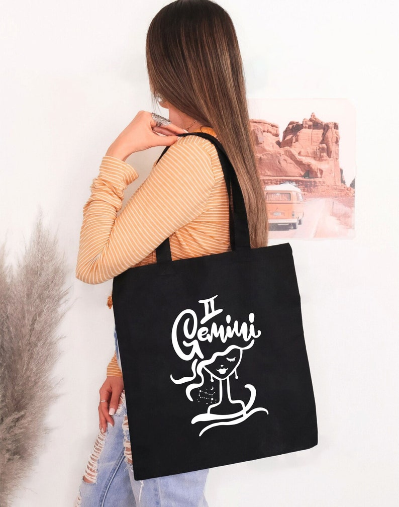 May include: A black tote bag with a white graphic of a woman's face and the text "Gemini" above it. The woman's face is stylized and has a constellation of stars around her head.