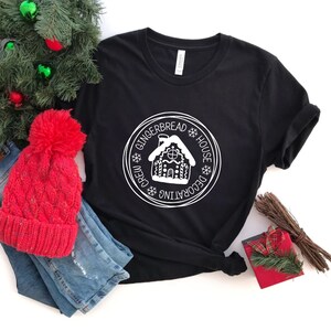 May include: A black short-sleeve t-shirt featuring a white circular graphic with a gingerbread house illustration in the center. The text reads GINGERBREAD HOUSE DECORATING CREW, accented by small snowflake icons. The shirt has a crew neckline.