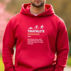 以下が含まれることがあります： 赤いフード付きスウェットシャツに、トライアスロンのシンボルと「TRIATHLETE」という文字の白いグラフィックが描かれています。その下に定義が書かれており、次のように書かれています。「（名詞）1つのスポーツが十分に難しいという事実を理解できない人。」