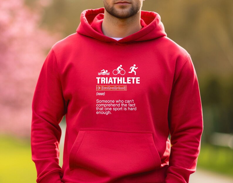 以下が含まれることがあります： 赤いフード付きスウェットシャツに、トライアスロンのシンボルと「TRIATHLETE」という文字の白いグラフィックが描かれています。その下に定義が書かれており、次のように書かれています。「（名詞）1つのスポーツが十分に難しいという事実を理解できない人。」