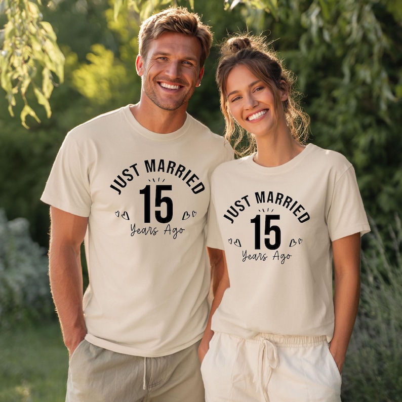 May include: Two people wearing matching beige t-shirts with the text "JUST MARRIED 15 Years Ago" in black. The shirts have a round neckline and short sleeves. The background is a blurred green outdoor setting.