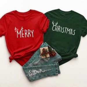 May include: Two Christmas-themed t-shirts, one red and one green, with white text. The red shirt says "MERRY" with antler and bow accents. The green shirt says "CHRISTMAS" with antler accents. Blue jeans and a gift box are in the image.