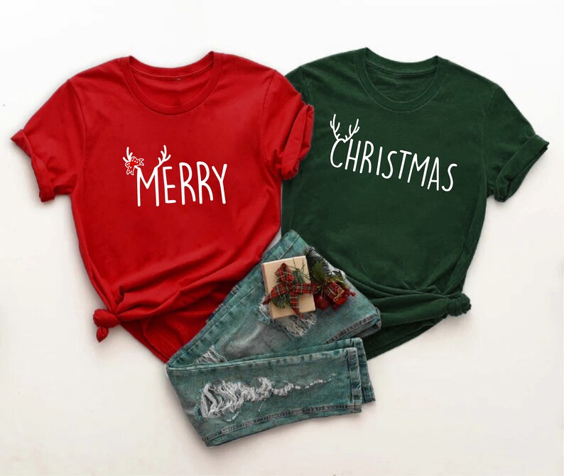 May include: Two Christmas-themed t-shirts, one red and one green, with white text. The red shirt says "MERRY" with antler and bow accents. The green shirt says "CHRISTMAS" with antler accents. Blue jeans and a gift box are in the image.