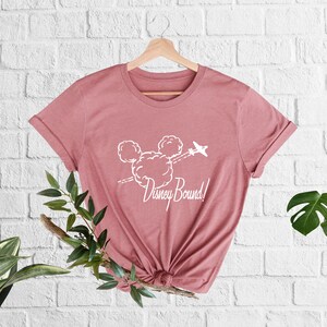 Peut inclure: Un t-shirt rose avec un graphique blanc repr&eacute;sentant la t&ecirc;te de Mickey Mouse faite de nuages avec un avion volant au-dessus. Le texte "Disney Bound!" est sous le graphique.