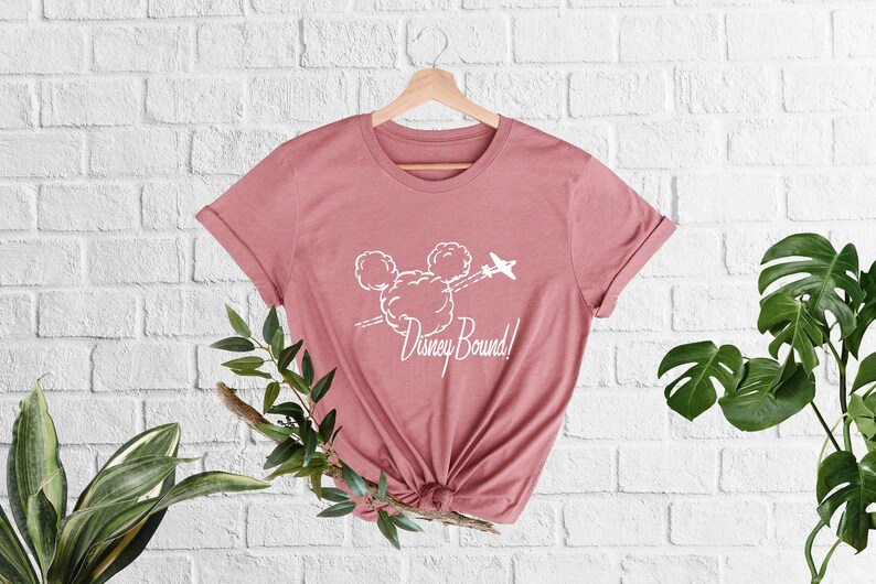 Peut inclure: Un t-shirt rose avec un graphique blanc repr&eacute;sentant la t&ecirc;te de Mickey Mouse faite de nuages avec un avion volant au-dessus. Le texte "Disney Bound!" est sous le graphique.