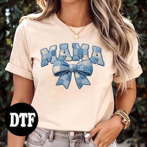 May include: Cream-colored t-shirt featuring the word "MAMA" in blue patchwork letters above a blue patchwork bow. The shirt has short sleeves and a round neckline. A black circle with the letters "DTF" is in the lower left corner.