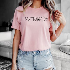 May include: A light pink t-shirt with the word "VIRGO" printed in black. The word is stylized with star and sun graphics. The shirt is paired with denim shorts.