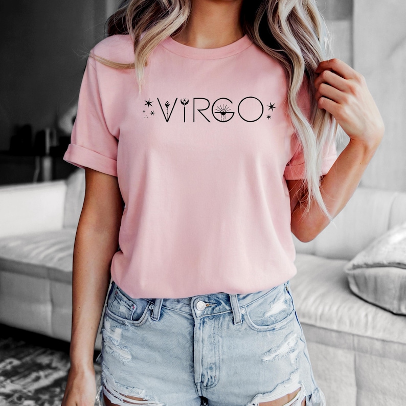 May include: A light pink t-shirt with the word "VIRGO" printed in black. The word is stylized with star and sun graphics. The shirt is paired with denim shorts.