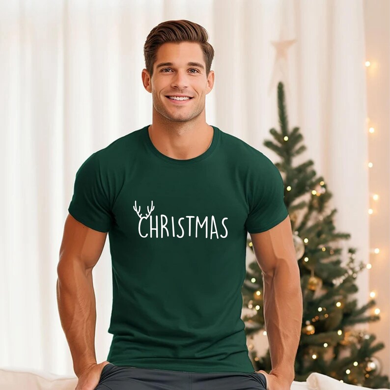 May include: A dark green t-shirt with the word "CHRISTMAS" in white script, featuring a deer antler design above the word. The shirt is short-sleeved and made of a soft material.