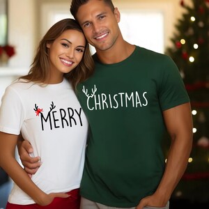 May include: A white t-shirt with the word "MERRY" and a red bow design, paired with a dark green t-shirt that says "CHRISTMAS" in white. Both shirts have a crew neck and short sleeves, perfect for the holiday season.