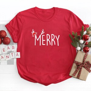 May include: A red t-shirt with the word "MERRY" in white script, adorned with antlers and a bow. The shirt is styled with Christmas ornaments, a wrapped gift, and the words "DEAR SANTA" spelled out in letter tiles, creating a festive holiday scene.