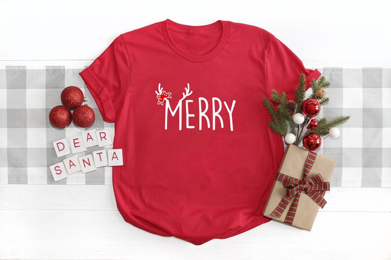 May include: A red t-shirt with the word "MERRY" in white script, adorned with antlers and a bow. The shirt is styled with Christmas ornaments, a wrapped gift, and the words "DEAR SANTA" spelled out in letter tiles, creating a festive holiday scene.