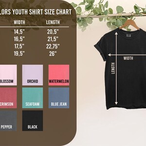 May include: Black Comfort Colors youth t-shirt hanging on a wooden hanger. The shirt's width is measured at 14.5 to 19.5 inches, and the length is 20.5 to 26 inches. A size chart with various colors is displayed.