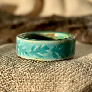 May include: A turquoise ceramic ring with a leaf pattern. The ring has a wide band and a glossy finish. The leaves are a lighter shade of turquoise, creating a contrast against the darker band. The ring is on a neutral-coloured fabric.