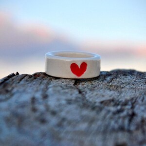 May include: A white ceramic ring with a hand-painted red heart design. The ring is a simple band style, perfect for everyday wear. The ring is shown on a weathered wooden surface.