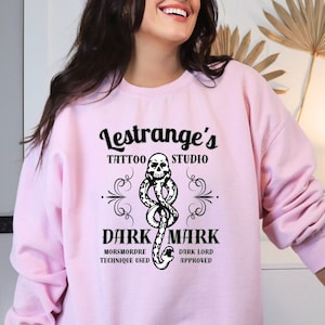 May include: Pink sweatshirt with a black graphic design that says "Lestrange's Tattoo Studio" with a skull and snake design. The text below says "Dark Mark" with the words "Morsmordre Technique Used" and "Dark Lord Approved" underneath.