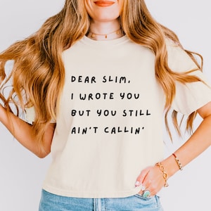 May include: A woman wearing a light yellow t-shirt with the text "Dear Slim, I wrote you but you still ain't callin'" printed in black.