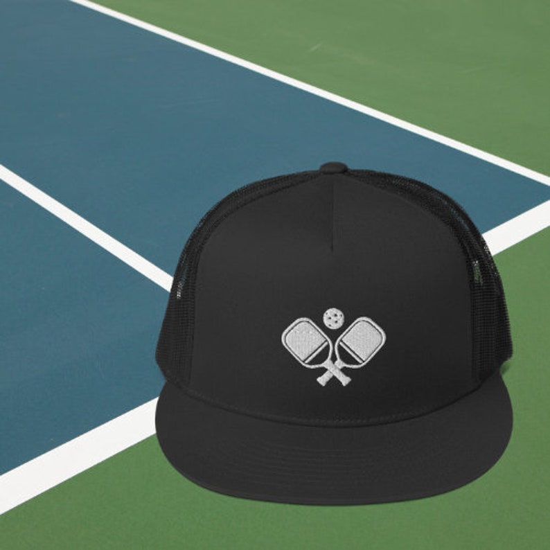 May include: A black mesh trucker hat with a white embroidered design of two crossed pickleball paddles and a ball. The hat has a black brim and a black mesh back.