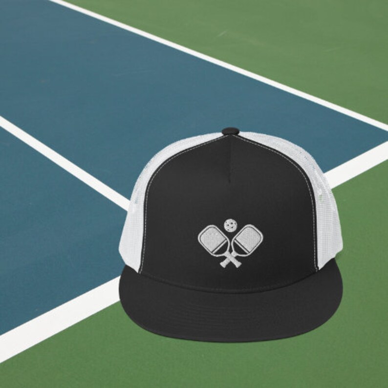 May include: A black and white trucker hat with a white embroidered design of two pickleball paddles and a ball. The hat has a black brim and a white mesh back.