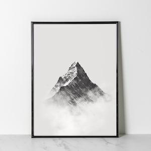 以下が含まれることがあります： 雲から現れる山の頂上を写した白黒写真。山はくっきりとピントが合っていますが、雲は柔らかくぼやけています。写真は黒い額縁に入っています。