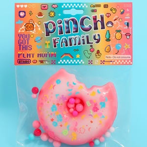 May include: A pink, bitten donut with colorful sprinkles and pink spheres, packaged in a clear plastic bag. The packaging features pixelated graphics and the words "Pinch Family" and "You Got This".