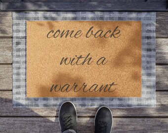 May include: A rectangular doormat with the text "come back with a warrant" in a cursive font. The mat has a tan center and a grey and white plaid border. Black shoes and blue jeans are visible.