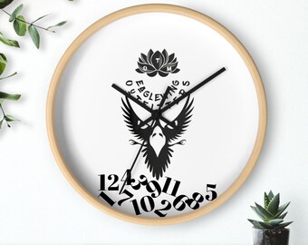 Puede incluir: Reloj de pared redondo con marco de madera clara y esfera blanca. El reloj presenta un gráfico negro de un pájaro con las alas extendidas y el texto "EAGLE OUTINGS". Los números negros están dispersos por la esfera.