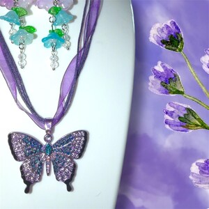 Op de afbeelding: Een paarse vlinderhangerketting met bijpassende oorbellen. De vlinderhanger is versierd met blauwe en paarse kristallen. De ketting heeft een paars lint. De oorbellen hebben blauwe en paarse bloembedels.