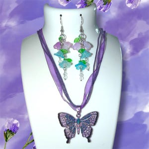 Op de afbeelding: Een sieradenset met een vlinderhangerketting en bijpassende oorbellen. De hanger heeft de vorm van een vlinder met blauwe en paarse strass-steentjes. De oorbellen hebben bloem- en bladbedels in blauwe, groene en paarse tinten.