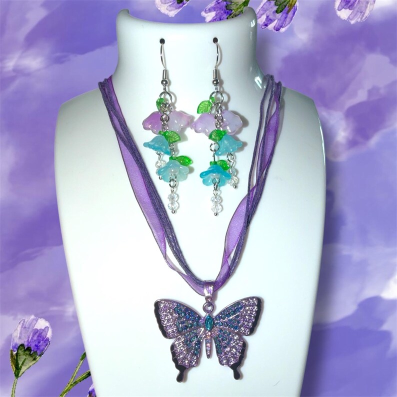 Op de afbeelding: Een sieradenset met een vlinderhangerketting en bijpassende oorbellen. De hanger heeft de vorm van een vlinder met blauwe en paarse strass-steentjes. De oorbellen hebben bloem- en bladbedels in blauwe, groene en paarse tinten.