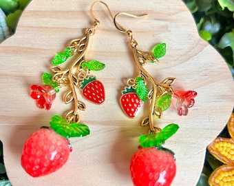 Op de afbeelding: Een paar goudkleurige oorhangers met een aardbeienthema. Elke oorbel heeft een gouden takontwerp met rode aardbeien, groene bladeren en rode en roze bloemaccenten. De oorbellen zijn geplaatst tegen een houten achtergrond.
