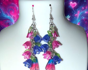 Op de afbeelding: Hangende oorbellen met clusters van kleine, doorschijnende roze en blauwe klokvormige bloembedels, groene bladaccenten en een enkele roze en blauwe vlindervleugelbedel met kleurverloop aan de onderkant, hangend aan zilverkleurige haken.