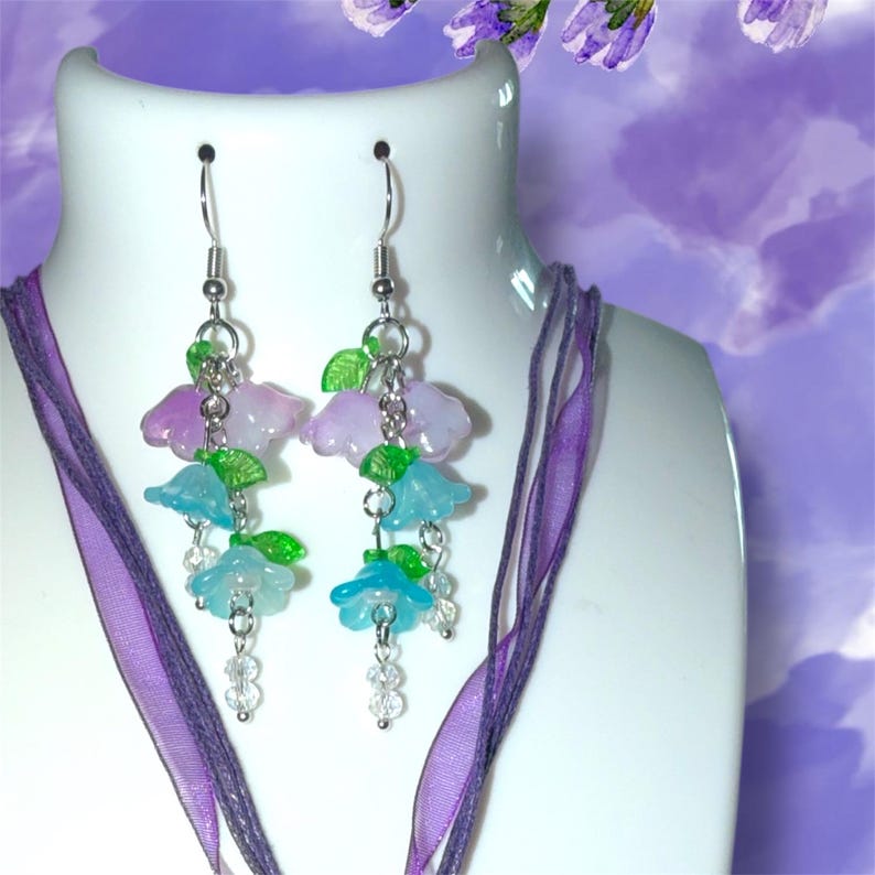 Op de afbeelding: Dangle oorbellen met clusters van doorschijnende blauwe en lavendelkleurige bloembedels, geaccentueerd met groene bladkralen en heldere kristallen kralen. De oorbellen hangen aan zilverkleurige haken. Een paars lint is gedrapeerd rond een witte mannequinhoofd.