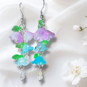 Op de afbeelding: Dangle oorbellen met doorschijnende blauwe, paarse en witte bloemvormige bedels met groene bladeren. De oorbellen hebben zilverkleurige haken en kettingen, met doorzichtige kralenhangers. Het bloemendesign is delicaat en grillig.
