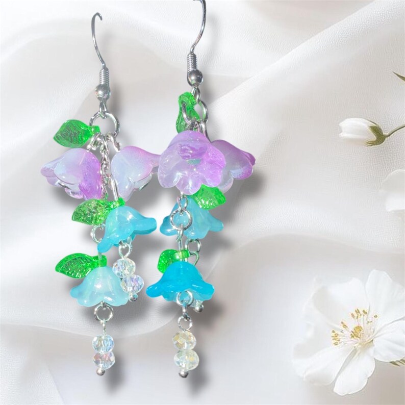 Op de afbeelding: Dangle oorbellen met doorschijnende blauwe, paarse en witte bloemvormige bedels met groene bladeren. De oorbellen hebben zilverkleurige haken en kettingen, met doorzichtige kralenhangers. Het bloemendesign is delicaat en grillig.