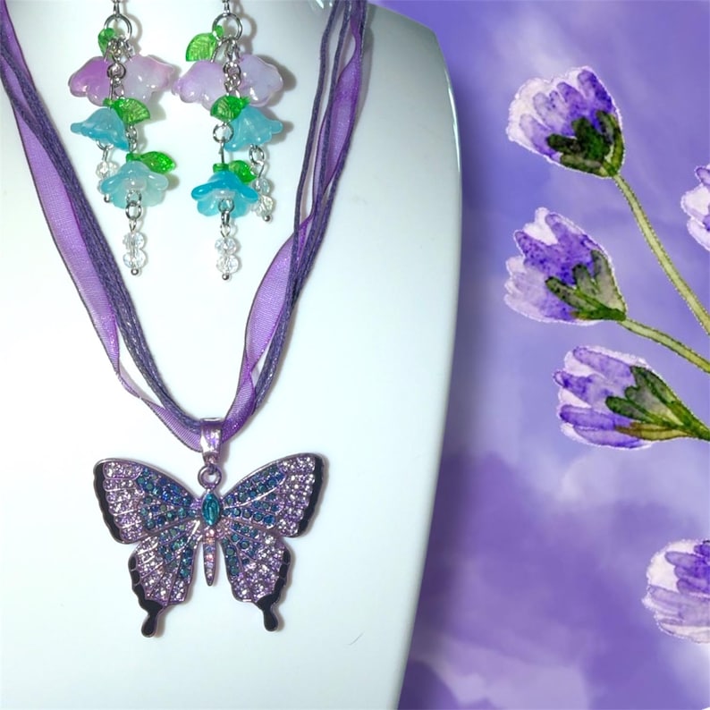 Op de afbeelding: Een paarse vlinderhangerketting met bijpassende oorbellen. De vlinderhanger is versierd met paarse en blauwe edelstenen, met een turquoise edelsteen in het midden. De oorbellen hebben bloemvormige kralen.
