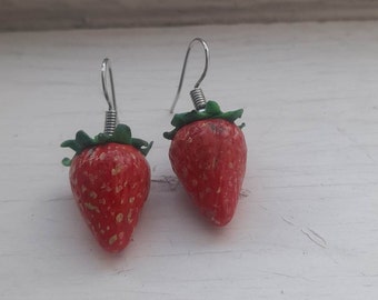 May include: A pair of dangle earrings featuring red and green strawberry charms. The earrings have silver hooks.