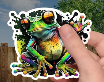 May include: A colorful sticker featuring a vibrant frog with rainbow hues. The frog has large, reflective eyes and is set against a black and green splatter design. The sticker is held against a backdrop of a wooden fence and green grass.