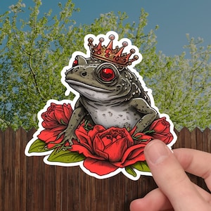 May include: A sticker featuring a cartoon frog wearing a gold crown and red eyes, surrounded by red roses. The frog is gray and green with a detailed design. The sticker is held in front of a wooden fence and greenery.