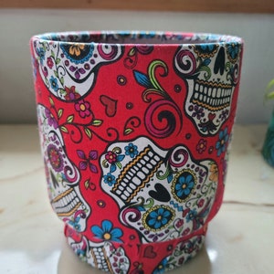 May include: A fabric-covered container with a vibrant red background. The container is adorned with a repeating pattern of colorful sugar skull designs, featuring white skulls with floral and decorative elements in blue, yellow, and pink.