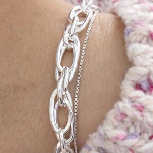 Peut inclure: Un bracelet argenté avec une chaîne à maillons ovales et épais, associé à un bracelet chaîne plus fin. Les bracelets sont portés sur un poignet, sur un fond doux et texturé. Les maillons de la chaîne sont polis.