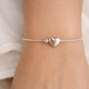 May include: A delicate silver bracelet with a heart-shaped charm and a small bead. The bracelet is made of a fine chain. The heart charm is the central design element. The bracelet is worn on a wrist.